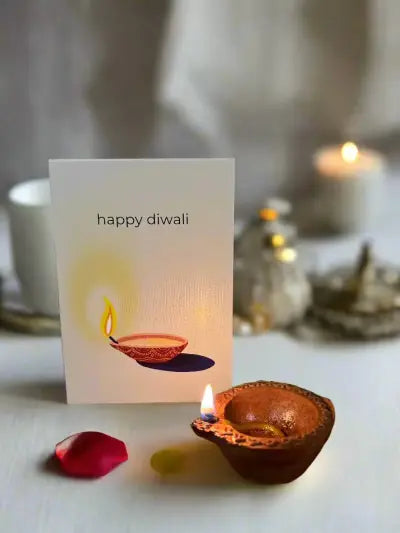 Diwali Greeting Card with artwork of a lit diya and text that says "Happy Diwali" pictured behind a real lit diya. 