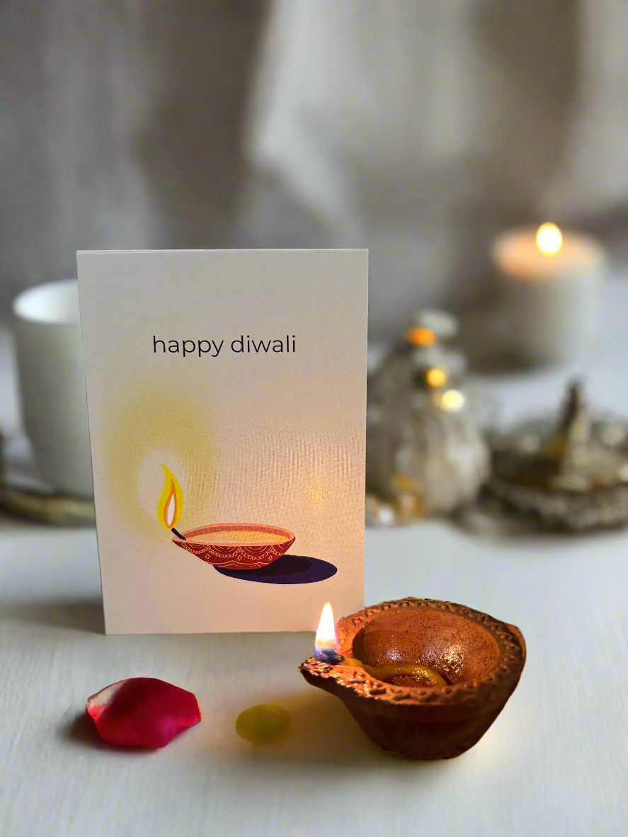 Diwali Greeting Card with artwork of a lit diya and text that says "Happy Diwali" pictured behind a real lit diya. 