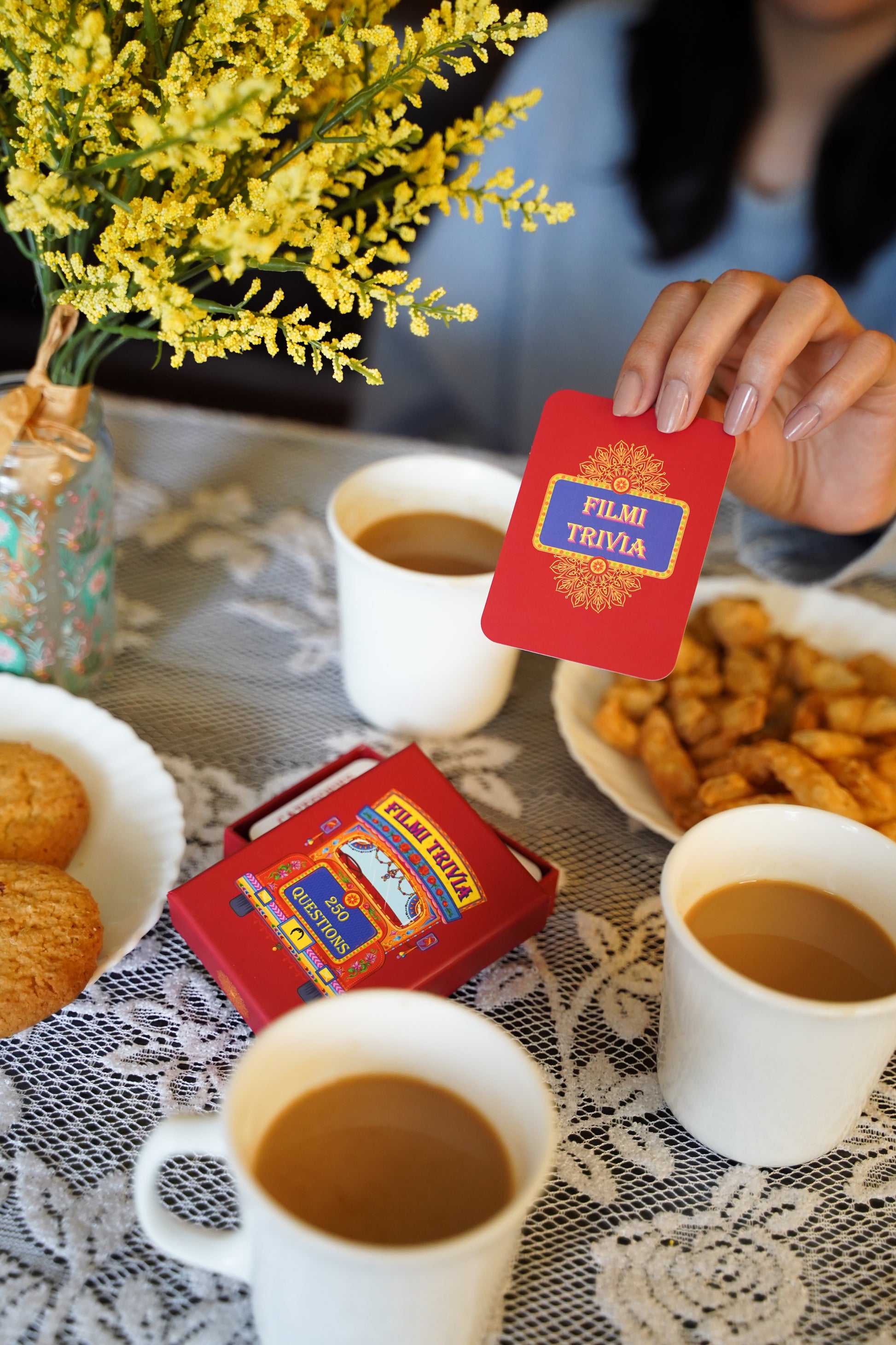 Person holding a 'Film Trivia' card over chai cups and snacks on a table.