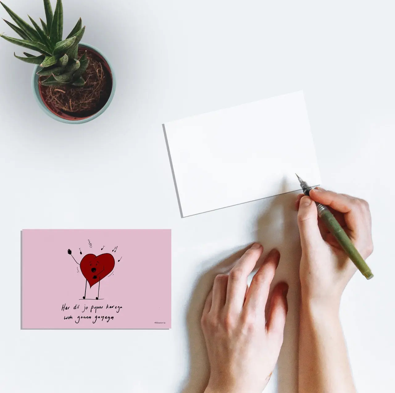 A person writing on a postcard with artwork of a heart singing and the text "Har dil jo pyaar karega woh gaana gayega" from the Bollywood film Har Dil Jo Pyar Karega (2000).