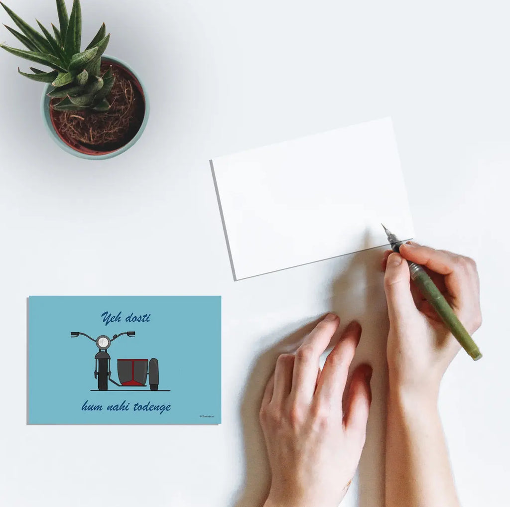 Person's hands writing a postcard featuring artwork of the famous motorcycle with a sidecar from the film Sholay featuring the text "Yeh dosti hum nahi todenge" in ode to the friendship between Jai and Veeru played by Amitabh Bachchan and Dharmendra.